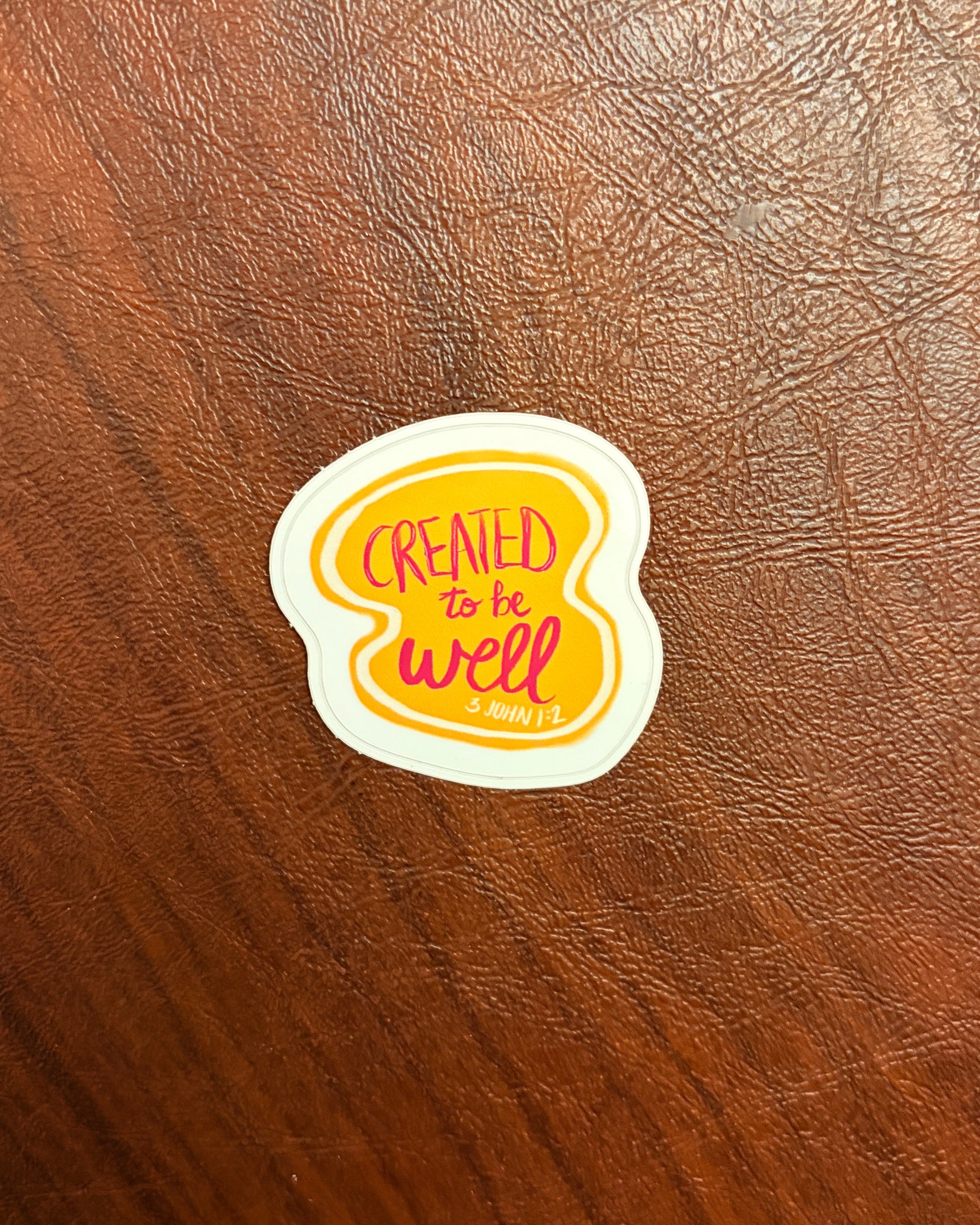 Sticker with 'Created to be well' text on a brown leather surface